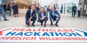 Drei Herren hocken vor dem Logo des Healthcare Hackathons auf dem Boden.