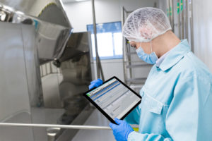 Mann mit Mundschutz, Haarnetz und Tablet vor einer Apparatur im Labor.