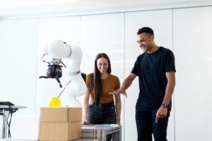 Junge Frau und junger Mann stehen in einem Raum vor einem weißen Roboterarm.