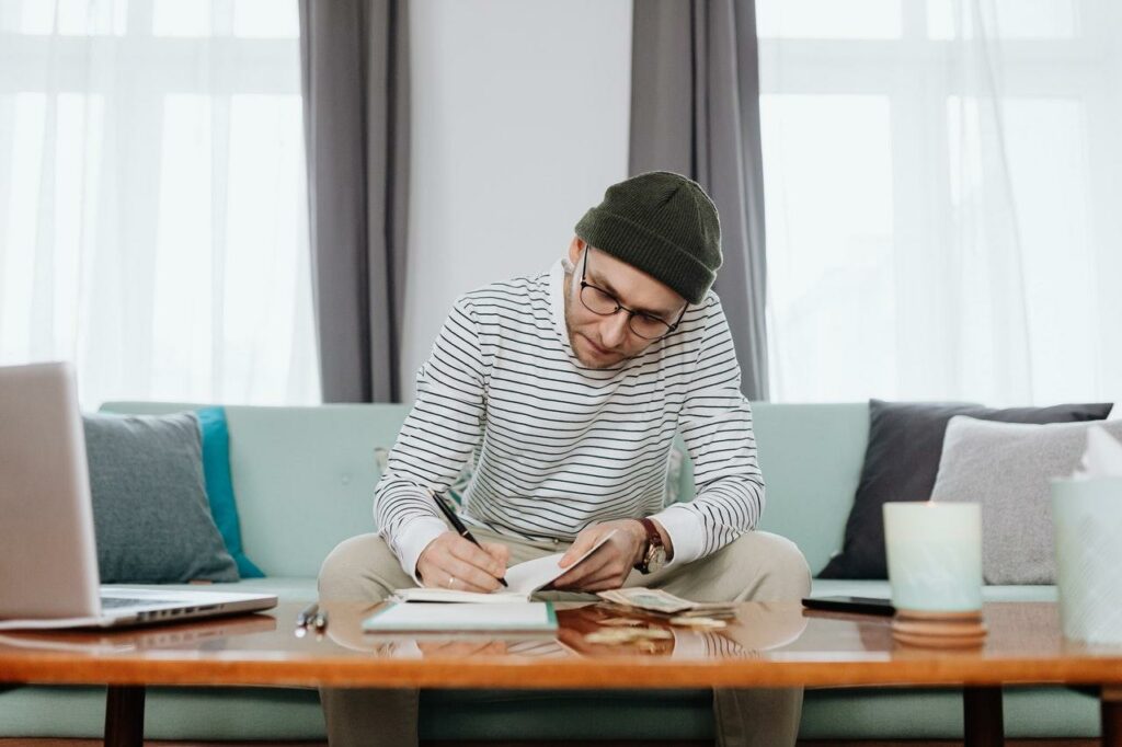 Junger Mann mit Mütze sitzt auf dem Sofa am Couchtisch und lernt oder arbeitet.