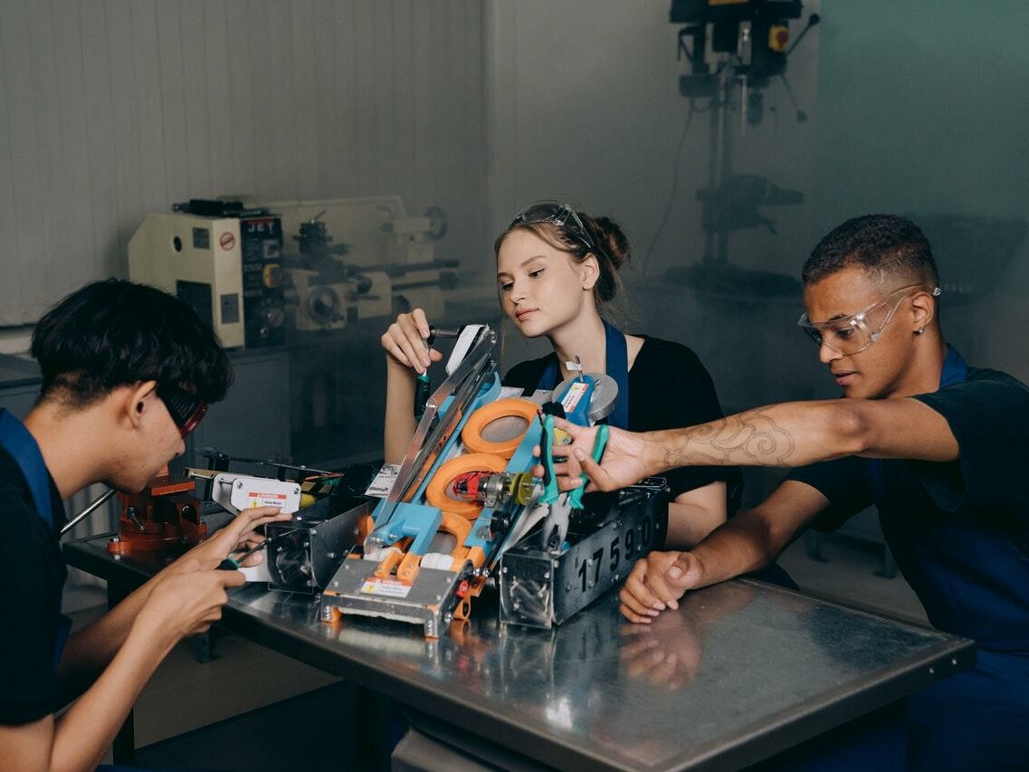 Eine junge Frau und zwei junge Männer sitzen am Tisch und beschäftigen sich mit ihrem Robotik-Projekt.