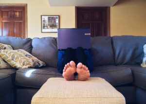 Eine Person sitzt auf dem Sofa. Gesicht wird vom Laptop verdeckt, man sieht nur die Füße.