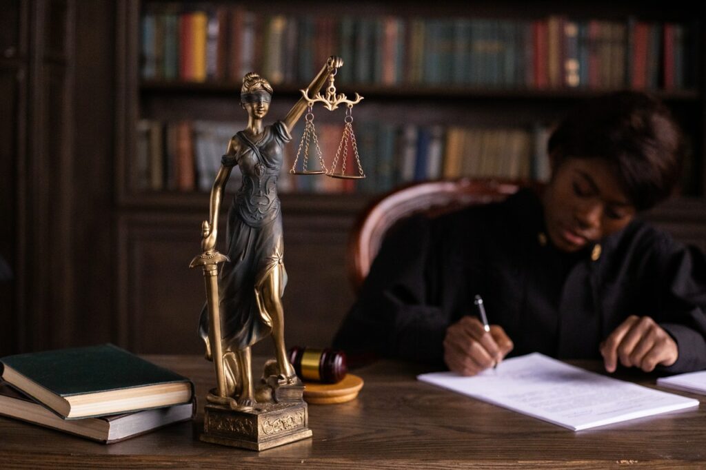 Eine Richterin sitzt an einem Tisch, vor sich die Justitia, und schreibt auf einem Blatt Papier.