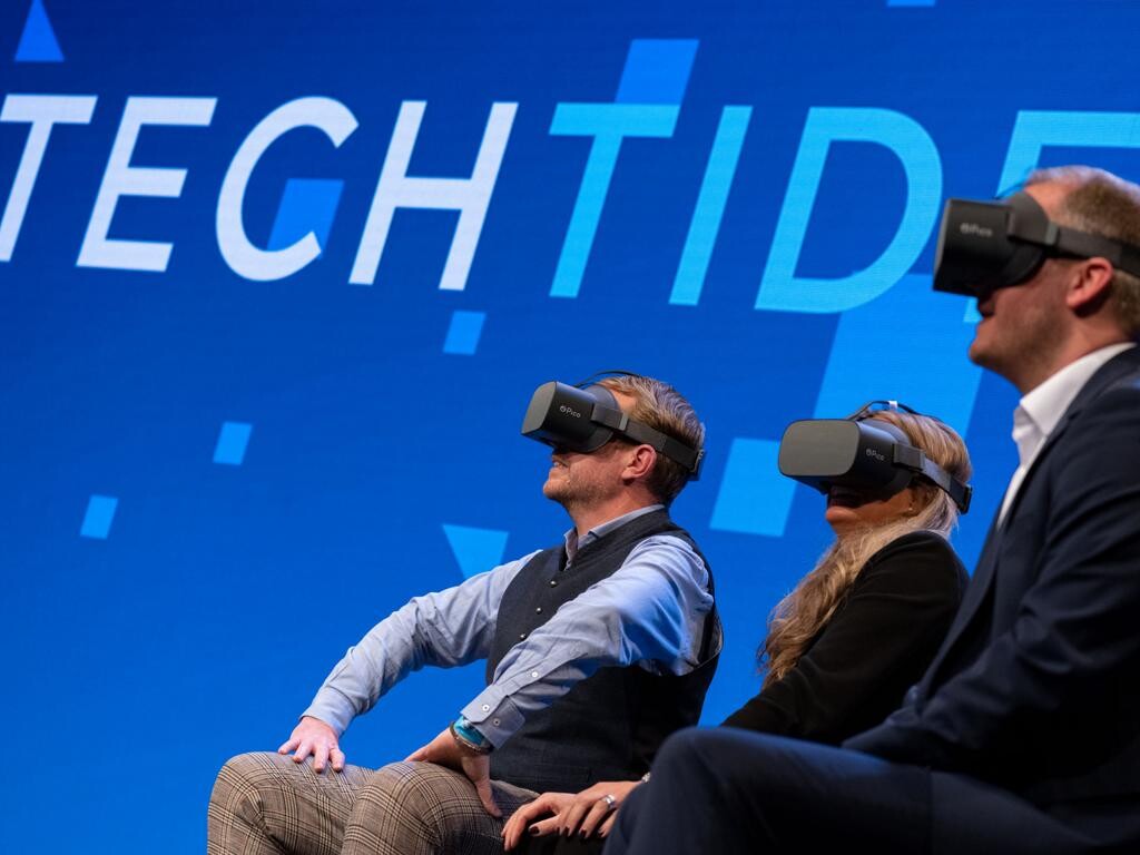 Drei Personen sitzen auf einer Bühne vor dem Techtide-Logo und haben jeweils eine VR-Brille auf.
