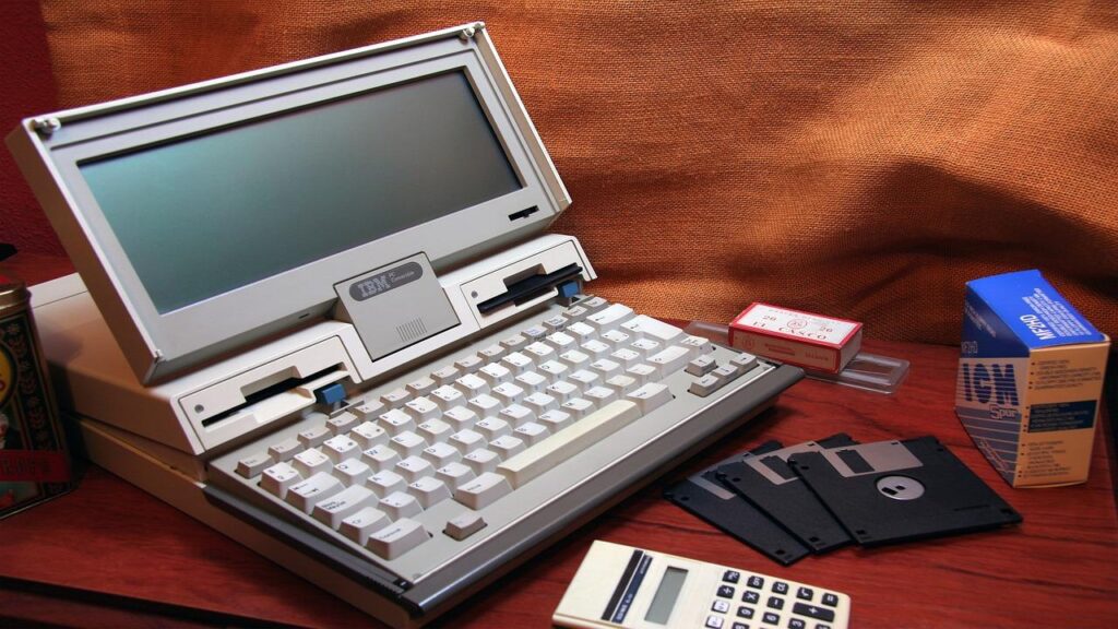 Alter IBM-Laptop neben Taschenrechner und Floppy-Disk auf einem Schreibtisch.