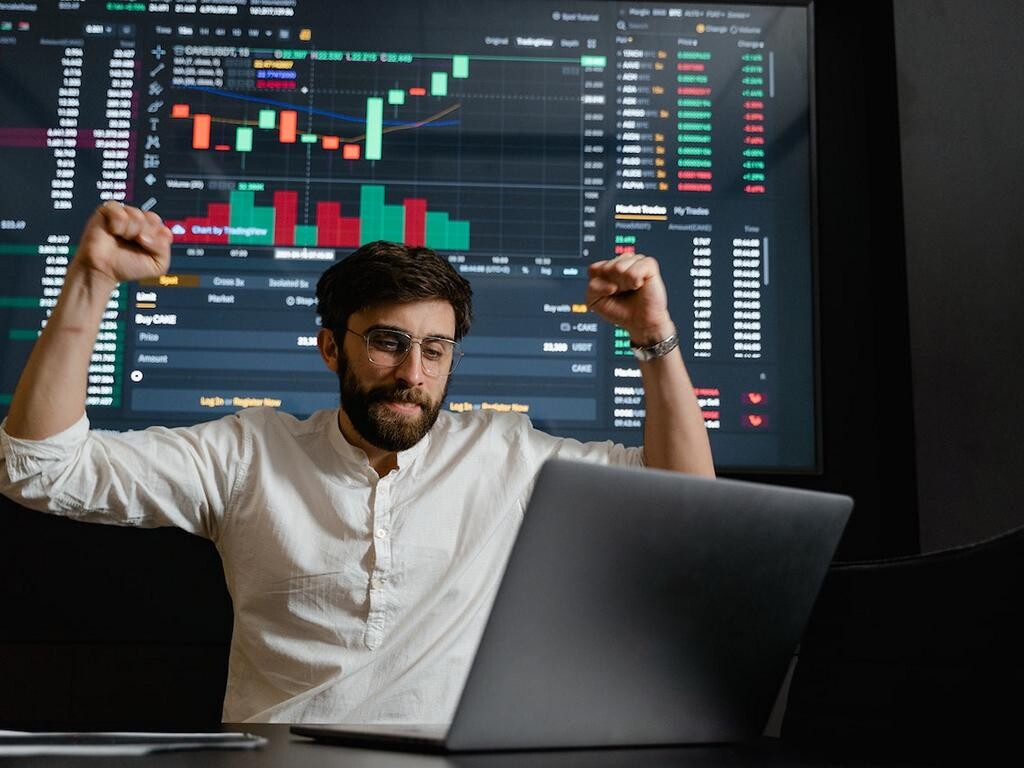 Ein Mann an einem Laptop und vor einem Bildschirm mit Börsenkursen reißt die Arme zum Jubeln nach oben.