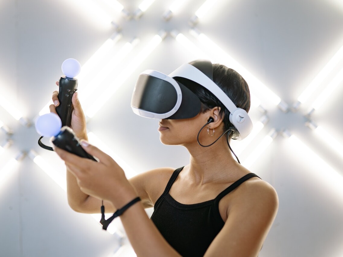 Eine Frau vor leuchtendem Hintergrund mit VR-Brille und zwei Handgeräten.