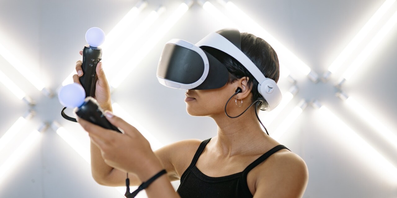 Eine Frau vor leuchtendem Hintergrund mit VR-Brille und zwei Handgeräten.