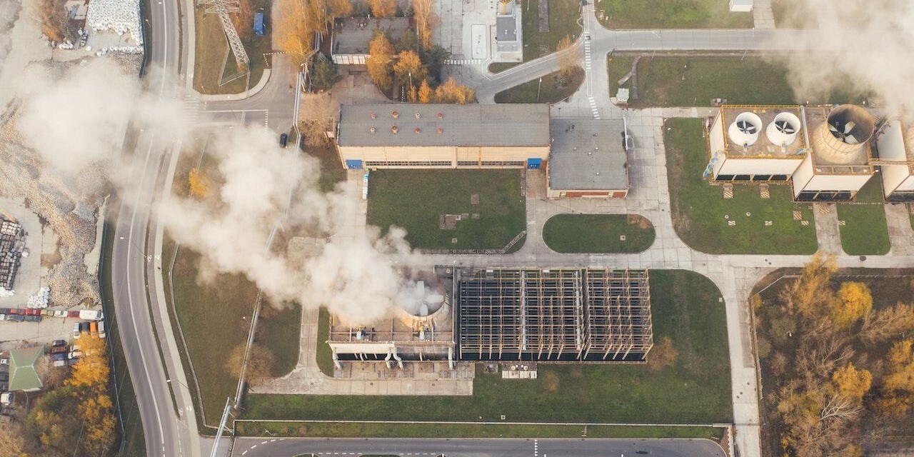 Luftbild einer Fabrik, aus deren Schornstein weißer Rauch aufsteigt.